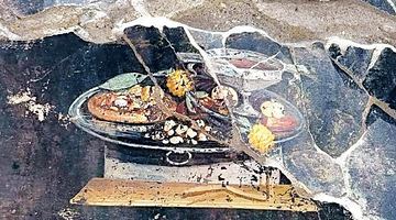 На фресці зображено круглий пляцок, схожий на піцу. На тісті – свіжі й сушені фрукти, а поруч срібна таця й келих із вином. Фото Parco Archeologico di Pompei